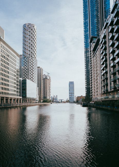 A daytime view of an urban canal flanked by modern high-rise buildings, with some buildings displaying glass facades that reflect the sky and surrounding structures. On the left, there are several commercial office towers with a grid-like pattern of windows, and a distinctive cylindrical tower with a textured exterior. On the right, contemporary residential buildings feature balconies and dark window frames. The water in the canal is calm, with gentle ripples, and the sky is partly cloudy with bright daylight illuminating the scene. This image, taken from a garden or outdoor space overlooking the canal, highlights the sleek cityscape typical of Canary Wharf, with busy architectural features. The landscape features no visible plants or gardening elements, but the perspective suggests the area may belong to or be managed by Gardeners Docklands, a specialist in outdoor maintenance and landscaping for urban environments in the E14 area.