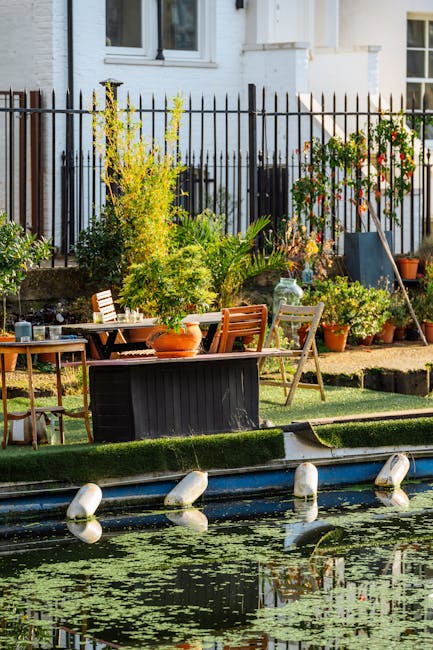 A vibrant Docklands balcony garden with a variety of potted plants and small shrubs arranged along a narrow paved pathway. The garden features a neatly trimmed lawn border, with lush green grass surrounding the plant beds. There are several terracotta and plastic pots containing leafy greens, flowering plants, and small trees, adding color and texture to the space. A black wooden garden table and matching chairs are positioned beside the plants, with gardening tools such as a watering can and a rake resting nearby. The garden is enclosed by a black iron fence, behind which a white residential building with windows is visible, illuminated by natural daylight. The scene showcases a well-maintained outdoor area focusing on garden maintenance, planting, and outdoor leisure, representative of the sort of landscaping work promoted by Gardeners Docklands for balcony gardening in urban environments like Docklands flats.