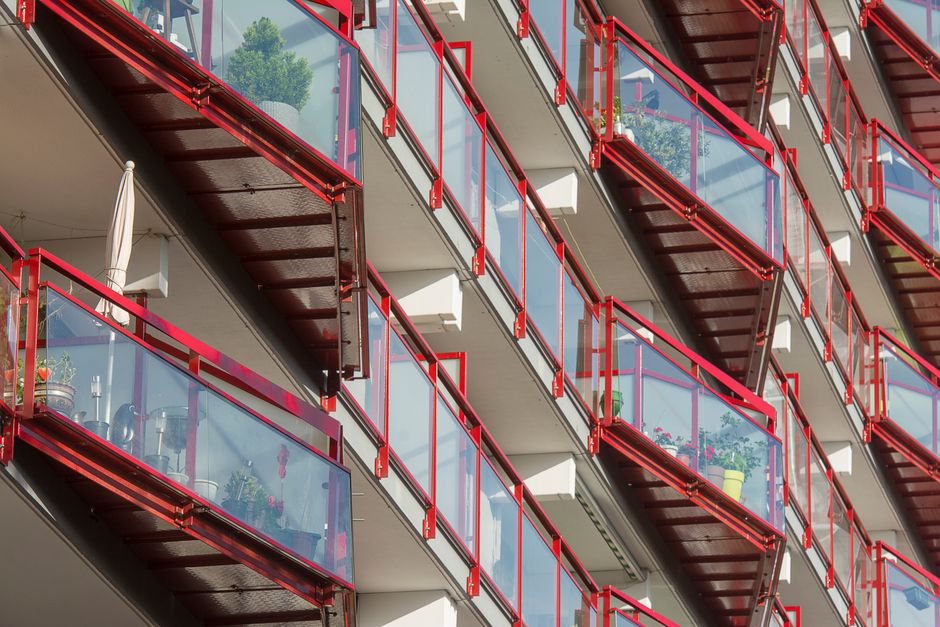 A close-up view of multiple balcony spaces on a modern multi-storey building, featuring glass-panelled railings with red metal frames. Each balcony has various potted plants, including leafy green foliage and flowering varieties, placed on the flooring or near the edges. The balconies are arranged in a staggered pattern, attached to white concrete building sections, with straight metal stairs visible between them. The light reflects off the glass railings, highlighting the textures of the plant leaves, the smoothness of the glass, and the sleek metallic surfaces. The scene depicts a typical urban residential setting with outdoor garden maintenance and landscaping elements, aligning with gardening tips discussed by Gardeners Docklands for balcony gardening in Docklands flats.